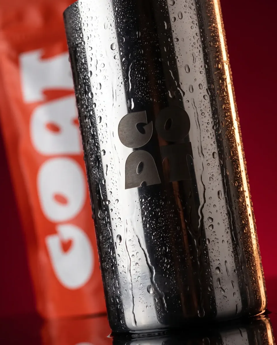 Copo metálico da Goat com gotas, com uma embalagem vermelha desfocada ao fundo